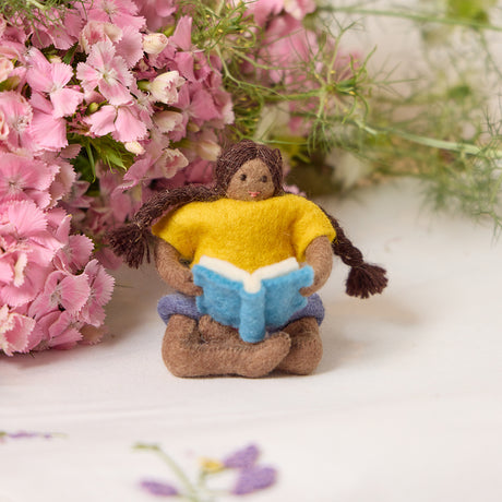 Handmade felt ornament of a young girl sitting cross-legged, absorbed in a book. Crafted from natural merino-blended wool, her focused gaze and relaxed posture capture the magic of reading. With braided brown hair and a warm-toned complexion, this ornament adds a cozy, literary touch to reading nooks, desks, or shelves. A perfect gift for bookworms, young readers, or anyone who treasures the quiet joy of a good story. Ready to hang with a string attached.