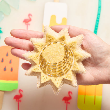 Natural wool sun charm ornament with golden embroidered details, embodying warmth and the promise of a new day.