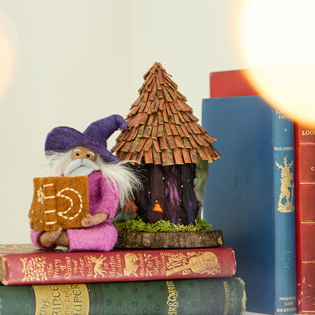Hand-felted wool ornament of a grey-bearded wizard wearing a pointed hat, holding an open spell-book beneath swirling stardust. Featuring a warm-toned complexion, white hair, and flowing robes, this whimsical figure is crafted from natural merino-blend wool. Includes a hanging string and measures approximately 5 inches tall.