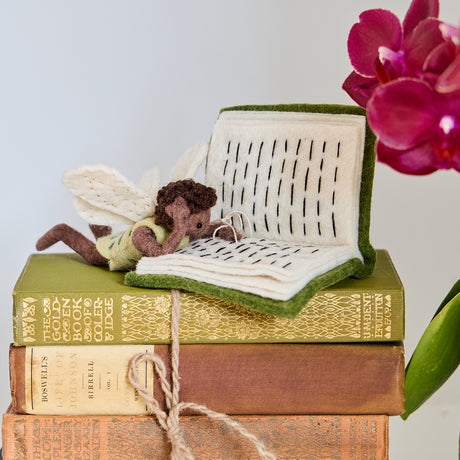 Handmade felt ornament of a fairy with delicate wings and a leafy green tunic, reading an oversized open book embroidered with whimsical details. Crafted from merino-blended wool, this limited-edition ornament is full of storybook charm. Perfect for fairy collectors, book lovers, and dreamers, it brings a magical, literary touch to bookshelves, holiday trees, or cozy reading spaces. Ready to hang with a string attached.