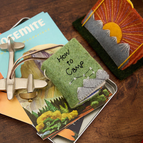 Hand-sewn felt ornament in the shape of a miniature book titled How to Camp, featuring embroidered pages with images of a tent, campfire, and fishing pole. Made from natural merino-blend wool, this whimsical ornament includes a hanging string and measures approximately 2.5 inches tall and 3 inches wide.