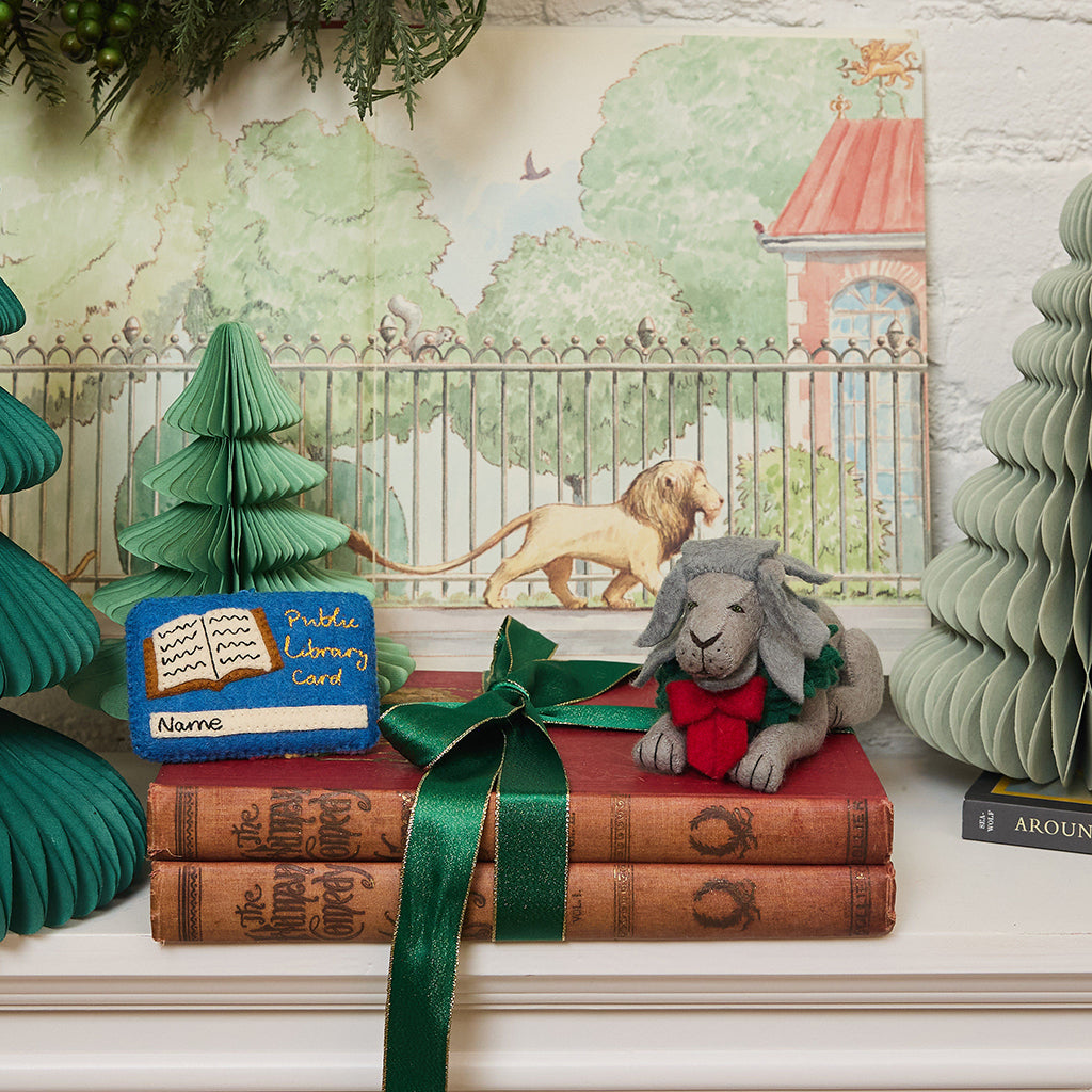 Handmade felt ornament of a regal lion statue adorned with a festive holiday wreath, inspired by the iconic lions outside the New York Public Library.  Shown with a library card ornament and stack of antique books. Crafted from merino-blended wool, this ornament captures the spirit of NYC, literary tradition, and holiday cheer. A thoughtful gift for book lovers, librarians, and New York enthusiasts, perfect for trees, bookshelves, or desks. Ready to hang with a string attached.