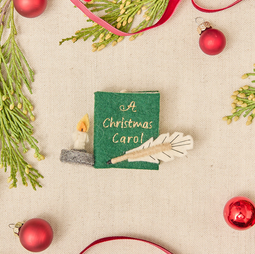 Miniature hand-felted wool ornament shaped like an open book titled A Christmas Carol, featuring embroidered details including a writing plume and a golden chamberstick. Crafted from merino-blend wool with a hanging string, the ornament evokes Victorian charm and the classic story of Ebenezer Scrooge. Measures approximately 2.25 inches high and 3.75 inches wide.