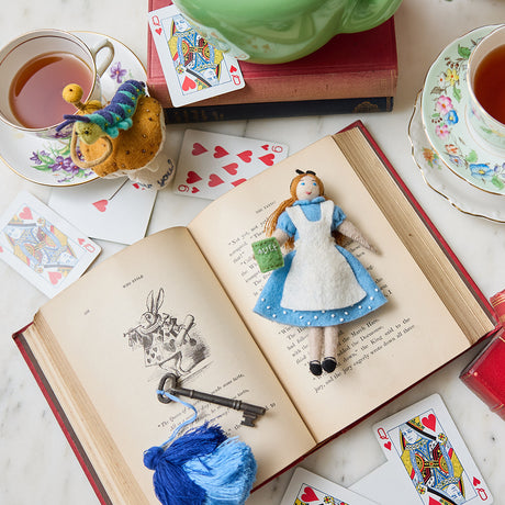 Hand-felted wool ornament of Alice from Alice in Wonderland, wearing her classic blue and white dress and holding a felt copy of Lewis Carroll's book. Crafted with embroidered details and made from merino-blend wool, the ornament includes a hanging string and measures approximately 5.75 inches tall