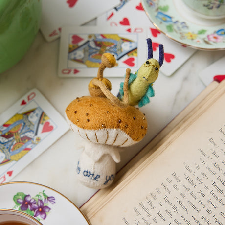 Hand-felted wool ornament of the Caterpillar from Alice in Wonderland, perched on a red-and-white mushroom with a hookah beside him. Features detailed embroidery and the phrase 'Who are you?' stitched at the base. Made from natural merino-blend wool, this whimsical ornament includes a hanging string and measures approximately 5.5 inches tall.