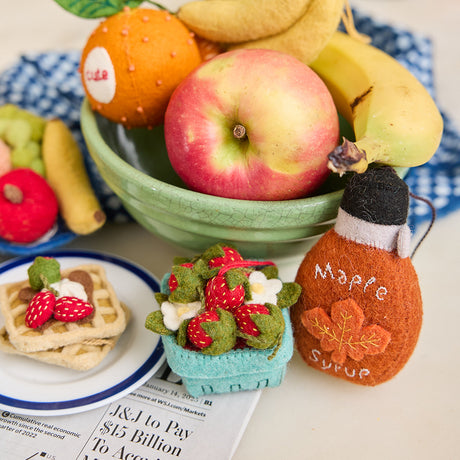 Felt ornament shaped like a stack of strawberry waffles, topped with three red strawberries, a swirl of white whipped cream, and a brown drizzle resembling Nutella. Handmade from soft wool felt with detailed embroidery, measuring approximately 2.5 inches tall and 1.5 inches wide, and comes with a hanging string. Pictured along with fresh fruit and ornaments of maple syrup and strawberries.