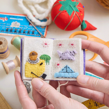 Hand-felted ornament shaped like a miniature crafting book, featuring three embroidered and appliquéd pages with illustrations of a pottery wheel, origami crane, knitting needles, and weaving loom. Made from merino-blend wool, this ornament includes a hanging string and measures approximately 2.5 inches tall. A charming tribute to handmade artistry and creative expression. GIF of pages of the book.