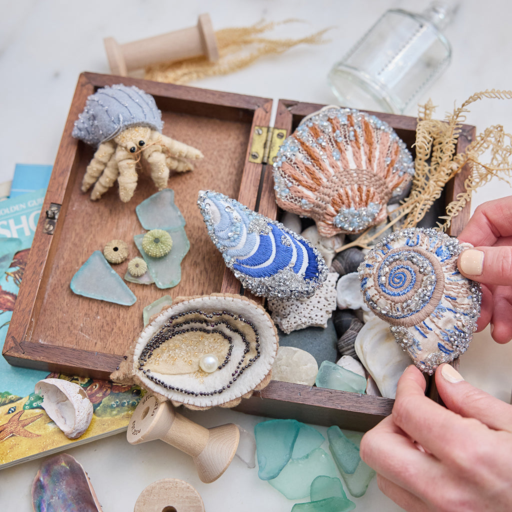 Craftspring’s Mussel Shell, Oyster, Moon Snail Shell, Scallop Shell, and Hermit Crab ornaments — all intricately beaded and embroidered, the shells made of satin and the hermit crab made of felt — displayed in a box of ocean treasures with seashells and sea glass, creating a playful and festive holiday scene, ethically crafted by artisans.