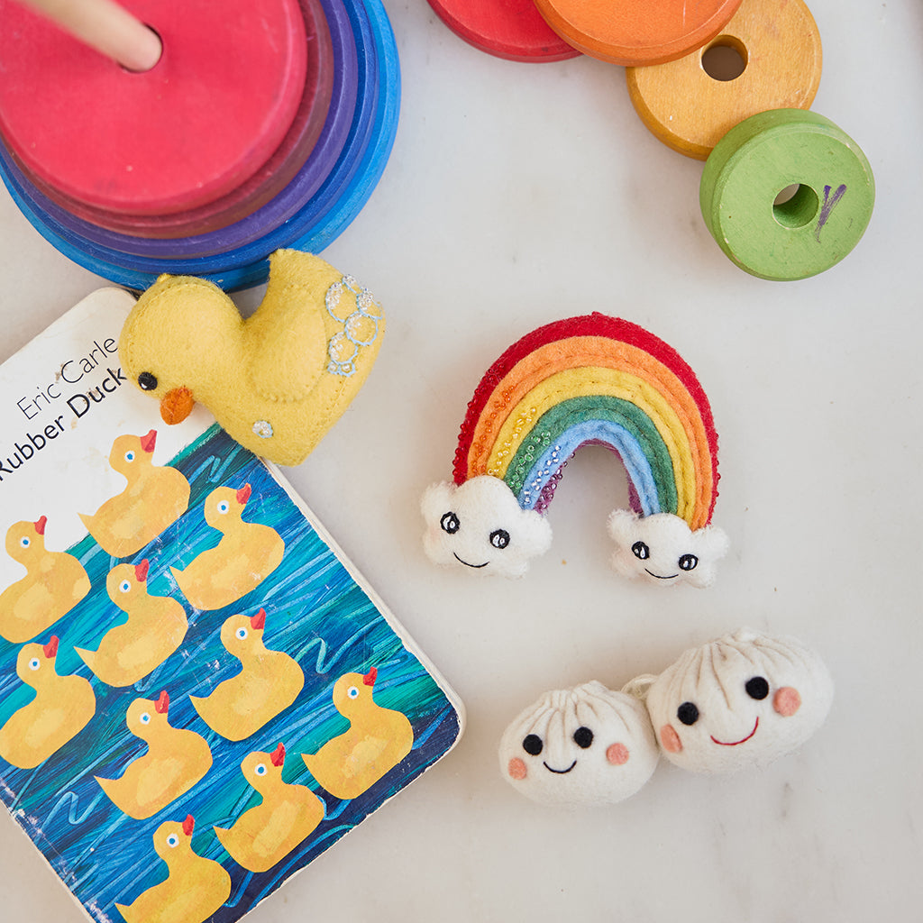 Handmade felt ornaments from Craftspring’s New Baby collection, featuring a Rubber Duck, Cute You & Me Rainbow, and smiling Dumplings—displayed together surrounded by children’s toys and a book, perfect for playful nursery decor and thoughtful baby shower gifting, ethically crafted by artisans using sustainable wool.