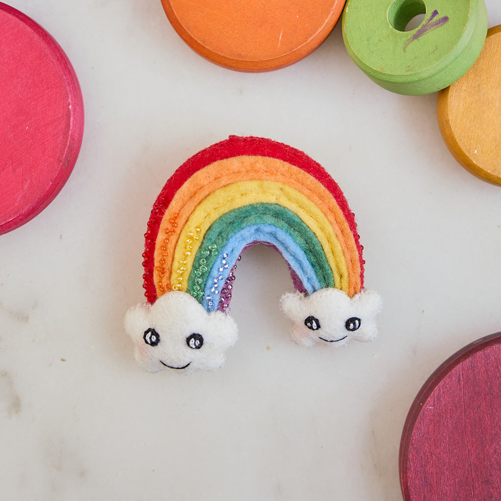 Handmade felt Cute You & Me Rainbow ornament from Craftspring, featuring intricate beaded and embroidered details with cute faces on clouds—displayed on marble surrounded by children’s toys, perfect for nursery decor and thoughtful baby shower gifting, ethically crafted by artisans using sustainable wool.
