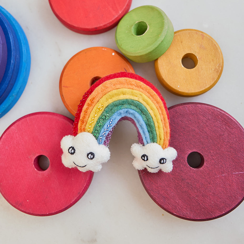 Handmade felt Cute You & Me Rainbow ornament from Craftspring, featuring intricate beaded and embroidered details with cute faces on clouds—displayed on marble surrounded by children’s toys, perfect for nursery decor and thoughtful baby shower gifting, ethically crafted by artisans using sustainable wool.