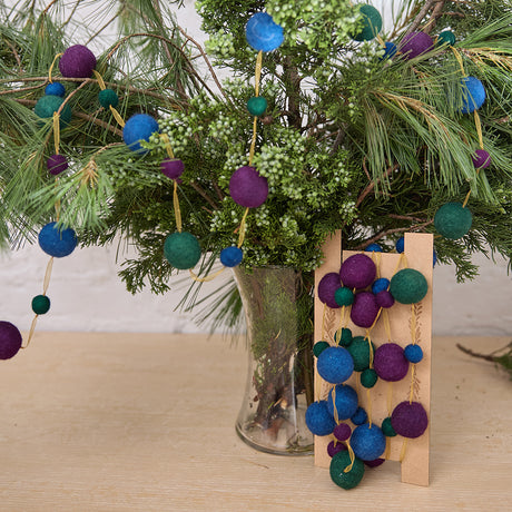 Handmade felt Jewel Tone Ball Garland from Craftspring, featuring deep jewel-tone felt balls strung on a gold ribbon—displayed among evergreen branches, perfect for adding rich, festive color and heirloom-quality holiday charm, ethically crafted by artisans using sustainable wool.