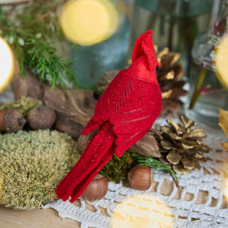 Craftspring’s handmade felt Cardinal ornament — featuring delicate beaded and embroidered details, displayed perched on a stick surrounded by acorns, moss, and pinecones for a cozy woodland holiday scene, perfect for Christmas decorating or gifting, ethically crafted by artisans using sustainable wool.