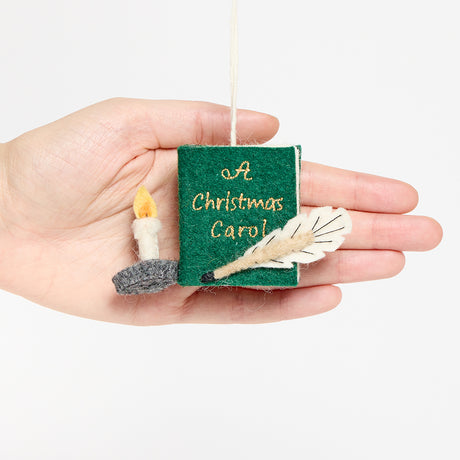 Miniature hand-felted wool ornament shaped like an open book titled A Christmas Carol, featuring embroidered details including a writing plume and a golden chamberstick. Crafted from merino-blend wool with a hanging string, the ornament evokes Victorian charm and the classic story of Ebenezer Scrooge. Measures approximately 2.25 inches high and 3.75 inches wide. Held in hand for scale.