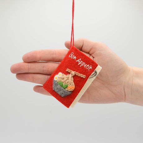 Miniature felt ornament shaped like a cookbook, hand-felted and embroidered with cooking-themed details including salt, pepper, vegetables, and a stovetop. Crafted from merino-blend wool with soft pages and a hanging string, this ornament measures approximately 2.5 inches tall. A cozy tribute to home cooking and culinary tradition. Held in a hand for scale reference.