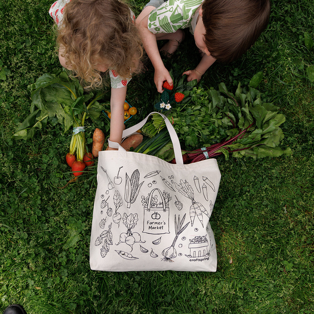Two children unpacking a Farmers Market tote bag filled with real produce and felt ornaments of fruits and vegetables evoke summertime berry picking and garden freshness. A sweet gift for foodies, kids, or lovers of handmade seasonal décor.