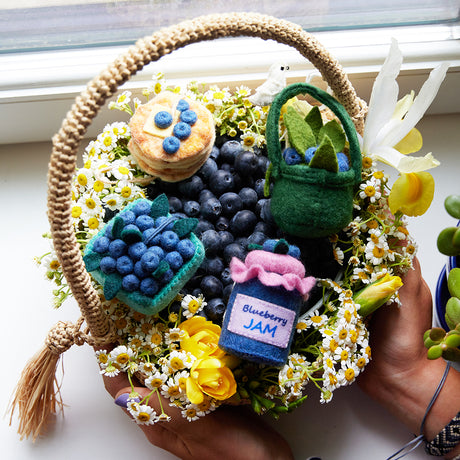 Hand-felted blueberry jam jar ornament with a gingham-style lid and a bunch of plump felt blueberries on top. Pictured in a spring gift basket of felt blueberry ornaments and flowers, a charming, summer-inspired gift for bakers and brunch lovers. Perfect for cottagecore décor, holiday trees, or breakfast-themed bundles