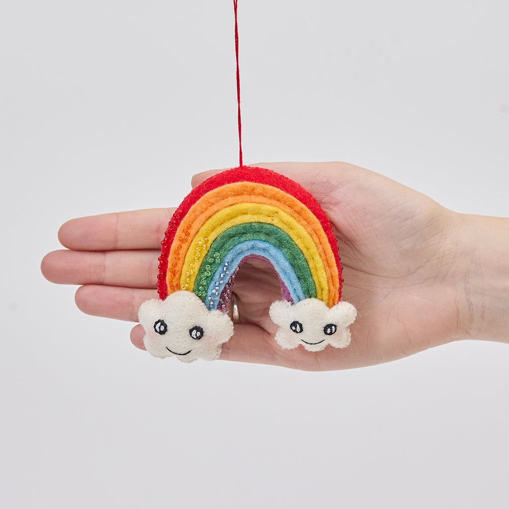 Handmade felt Cute You & Me Rainbow ornament from Craftspring held in front of a hand to show size—featuring intricate beaded and embroidered details with cute faces on clouds, ethically crafted by artisans using sustainable wool.
