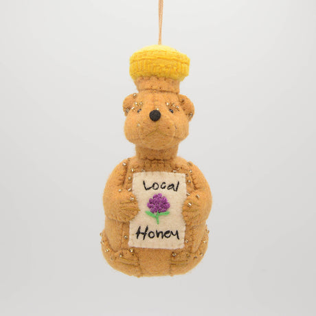 Festive hand-felted wool ornament of a honey bear jar, sparkling with beaded details and a stitched clover on its chest. Inspired by Winnie the Pooh, this cozy golden bear makes a charming holiday gift and nostalgic keepsake. Includes a hanging string and measures approximately 4.5 inches tall — perfect for tree decorating, gift-topping, or stocking stuffing.