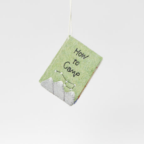 Hand-sewn felt ornament in the shape of a miniature book titled How to Camp, featuring embroidered pages with images of a tent, campfire, and fishing pole. Made from natural merino-blend wool, this whimsical ornament includes a hanging string and measures approximately 2.5 inches tall and 3 inches wide.