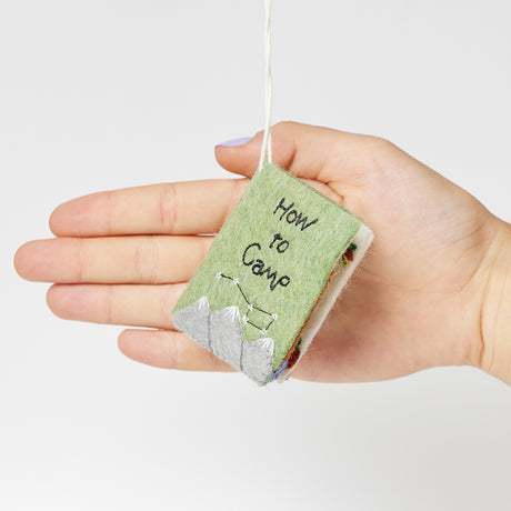 Hand-sewn felt ornament in the shape of a miniature book titled How to Camp, featuring embroidered pages with images of a tent, campfire, and fishing pole. Made from natural merino-blend wool, this whimsical ornament includes a hanging string and measures approximately 2.5 inches tall and 3 inches wide. With a hand for scale.