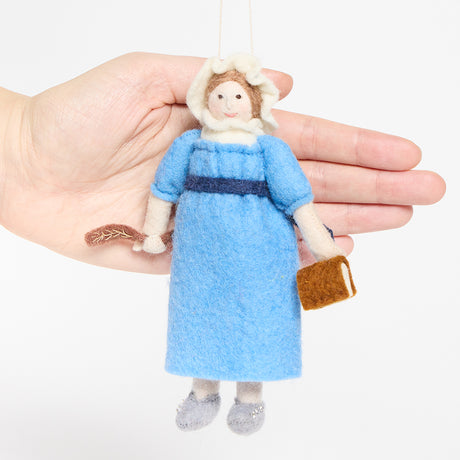 Handcrafted felt ornament of Jane Austen, featuring the beloved author standing with a feather quill in one hand and a felt copy of Pride and Prejudice in the other. She wears a period-style dress and bonnet, all made from natural merino-blend wool with fine stitched details. Includes a string for hanging and measures approximately 6 inches tall. Hand for scale reference.