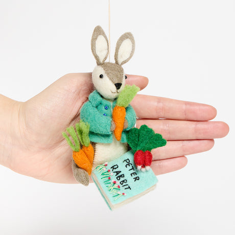 Handmade wool ornament of Peter Rabbit, featuring the beloved children's book character sitting with a carrot in one paw and an embroidered book on his lap. He wears his signature blue jacket, crafted with detailed stitching. Made from natural felted merino-blend wool, the ornament is ready to hang and sized approximately 4.5 inches tall. With a hand for scale