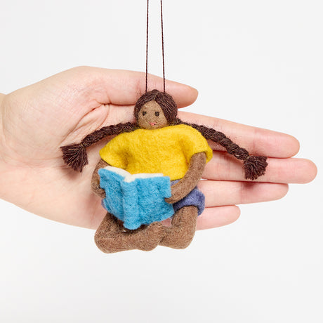 Handmade felt ornament of a young girl sitting cross-legged, absorbed in a book. Crafted from natural merino-blended wool, her focused gaze and relaxed posture capture the magic of reading. With braided brown hair and a warm-toned complexion, this ornament adds a cozy, literary touch to reading nooks, desks, or shelves. A perfect gift for bookworms, young readers, or anyone who treasures the quiet joy of a good story. Ready to hang with a string attached.