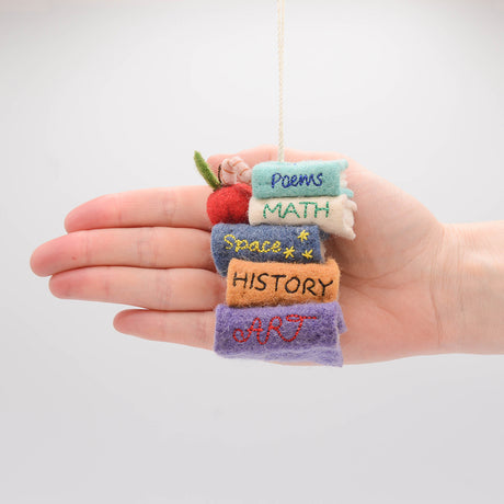 Handmade felt ornament featuring a miniature stack of colorful books, meticulously crafted from natural wool using traditional felting techniques. This eco-friendly and lightweight ornament adds a cozy, literary touch to holiday trees, bookshelves, or reading nooks. A charming gift for bookworms, teachers, and students, perfect for any occasion or year-round decor. Hand in photo for scale reference.