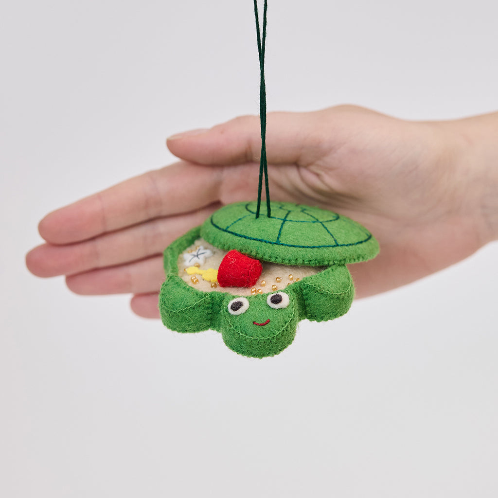 Handmade felt Turtle Sandbox ornament from Craftspring held in front of a hand to show its size—with the turtle shell cover slightly open revealing beaded sand, a bucket, mini shovel, and starfish inside, ethically crafted by artisans in India using sustainable wool.