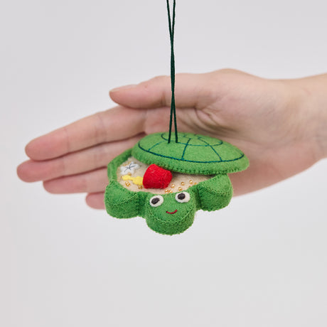 Handmade felt Turtle Sandbox ornament from Craftspring held in front of a hand to show its size—with the turtle shell cover slightly open revealing beaded sand, a bucket, mini shovel, and starfish inside, ethically crafted by artisans in India using sustainable wool.
