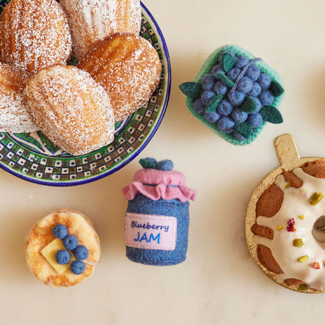 Hand-felted blueberry jam jar ornament with a gingham-style lid and a bunch of plump felt blueberries on top.  Alongside brunch favorites, both real and made of felt like pancakes and a pint of blueberries. Made from merino-blend wool, this 3-inch tall ornament is a charming, summer-inspired gift for bakers and brunch lovers. Perfect for cottagecore décor, holiday trees, or breakfast-themed bundles.