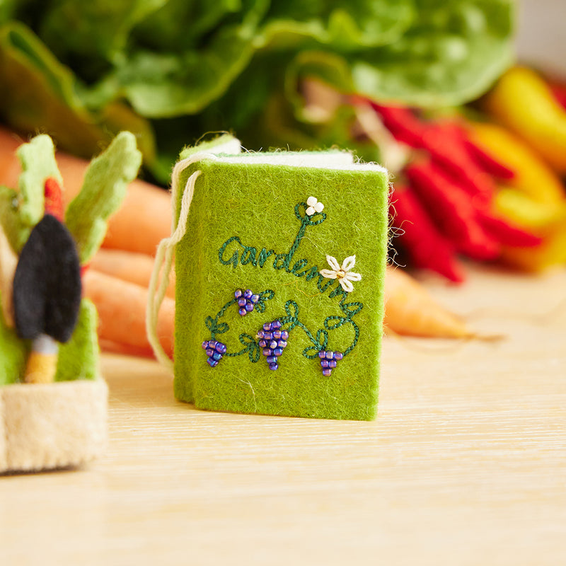 Handcrafted felt ornament shaped like a miniature gardening book, featuring embroidered and appliqué illustrations of seeds, flowers, and vegetables on its open pages. Made from merino-blend wool in vibrant colors, this ornament includes a hanging string and measures approximately 2.5 inches tall. A whimsical tribute to gardening and nature lovers.
