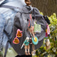 Hand-sewn felt ornaments hanging
 from a backpack including miniature book titled How to Camp, featuring embroidered pages with images of a tent, campfire, and fishing pole. Made from natural merino-blend wool, this whimsical ornament includes a hanging string and measures approximately 2.5 inches tall and 3 inches wide.