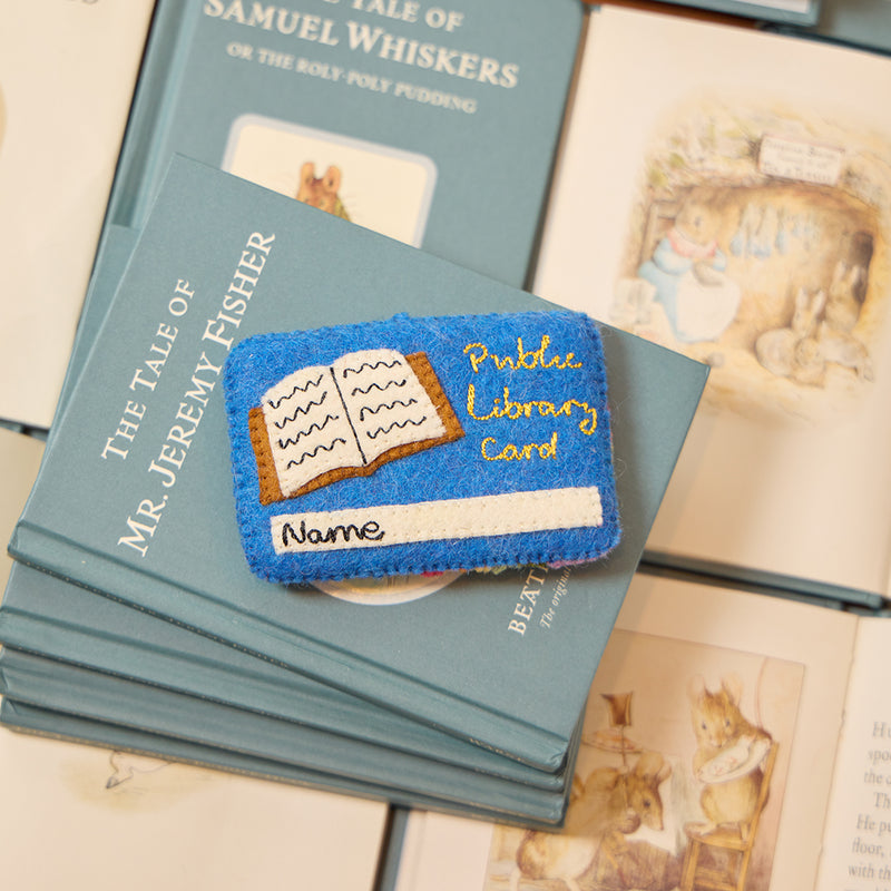 Handmade felt ornament shaped like a vintage library card, featuring intricate hand embroidery. One side displays a classic library card with a name field and book icon, while the reverse showcases a colorful rainbow, lightbulb, and whimsical scanning barcode. Crafted from soft merino-blended wool, this ornament celebrates libraries, learning, and the joy of reading. Perfect for bookworms, librarians, and teachers, it adds a literary touch to trees, reading corners, and classrooms.