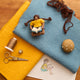 Handmade felt ornament of a young girl sitting cross-legged, absorbed in a book with the design sketch and materials. Crafted from natural merino-blended wool, her focused gaze and relaxed posture capture the magic of reading. With braided brown hair and a warm-toned complexion, this ornament adds a cozy, literary touch to reading nooks, desks, or shelves. A perfect gift for bookworms, young readers, or anyone who treasures the quiet joy of a good story. Ready to hang with a string attached.