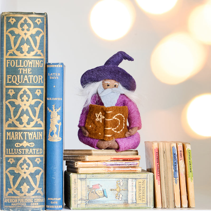 Hand-felted wool ornament of a grey-bearded wizard wearing a pointed hat, holding an open spell-book beneath swirling stardust. Featuring a warm-toned complexion, white hair, and flowing robes, this whimsical figure is crafted from natural merino-blend wool. Includes a hanging string and measures approximately 5 inches tall.