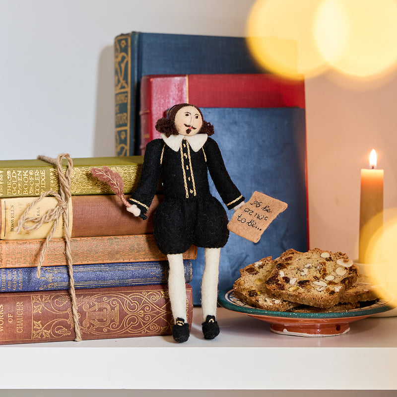 Hand-felted wool ornament of William Shakespeare, featuring the playwright in a classic doublet and hose with a feathered cap and a felt manuscript in hand. Crafted from natural merino-blend wool, the ornament captures his expressive posture and literary charm. Includes a string for hanging and measures approximately 7.5 inches tall.