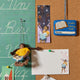 Handmade felt ornament of a young girl sitting cross-legged, absorbed in a book. Nearby are a library card and bookmark and note card.Crafted from natural merino-blended wool, her focused gaze and relaxed posture capture the magic of reading. With braided brown hair and a warm-toned complexion, this ornament adds a cozy, literary touch to reading nooks, desks, or shelves. A perfect gift for bookworms, young readers, or anyone who treasures the quiet joy of a good story. Ready to hang with a string attached.