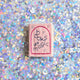 Hot pink felt ornament shaped like a small book, featuring delicately embroidered open pages. Inspired by the beloved Brooklyn bookstore Books Are Magic, this hand-felted wool ornament includes a hanging string and measures approximately 2.5 inches tall. A colorful tribute to the joy of reading and storytelling