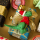 Handmade felt Santasaurus T-Rex ornament from Craftspring, displayed on top of a festive wrapped present—featuring a red Santa hat, red sweater, and embroidered details, perfect for dino-loving kids and joyful holiday gifting, ethically crafted using sustainable wool.