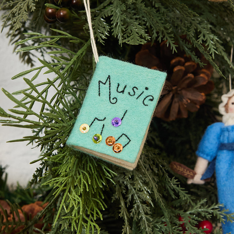 Handcrafted felt ornament shaped like a miniature music book, featuring three pages with embroidered musical notes, instruments, and a ballerina in mid-twirl. Made from merino-blend wool with appliqué details, this ornament includes a hanging string and measures approximately 2.5 inches tall by 3 inches wide. A joyful tribute to music, dance, and artistic expression.