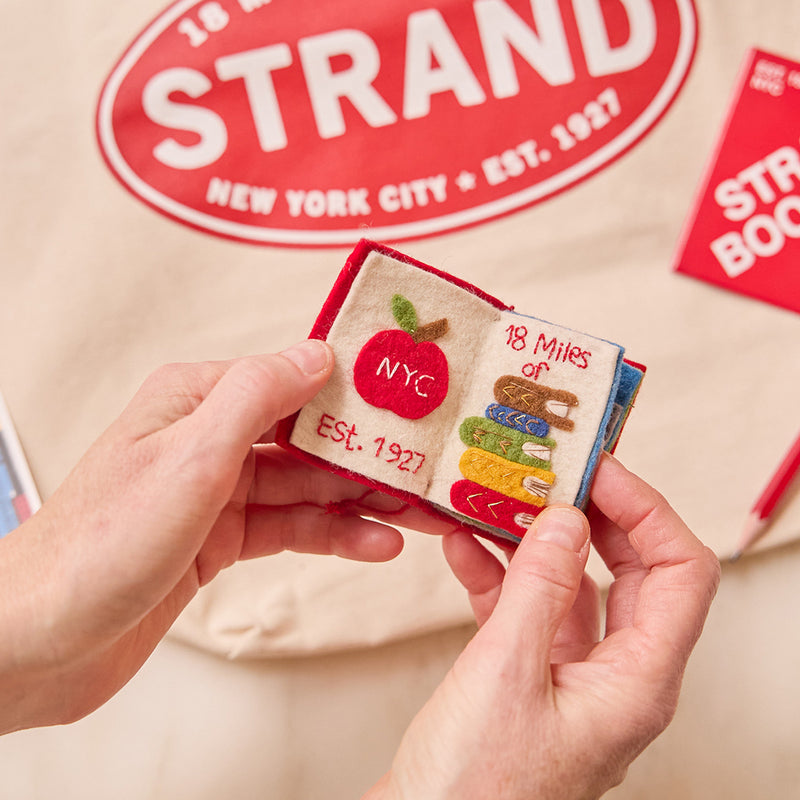 Handmade felt ornament shaped like a miniature book, featuring a red cover with the classic Strand Bookstore logo. The book opens to reveal three pages with whimsical, hand-stitched NYC scenes, including yellow taxis, skyscrapers, and pigeons with pizza. Crafted from felted wool, this ornament celebrates the magic of New York City and The Strand, making it a perfect gift for NYC enthusiasts, book lovers, and Strand fans. Ready to hang with a string attached.