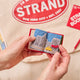 Handmade felt ornament shaped like a miniature book, featuring a red cover with the classic Strand Bookstore logo. The book opens to reveal three pages with whimsical, hand-stitched NYC scenes, including yellow taxis, skyscrapers, and pigeons with pizza. Crafted from felted wool, this ornament celebrates the magic of New York City and The Strand, making it a perfect gift for NYC enthusiasts, book lovers, and Strand fans. Ready to hang with a string attached. Open to page of NYC street with taxi and Stand 