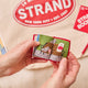Handmade felt ornament shaped like a miniature book, featuring a red cover with the classic Strand Bookstore logo. The book opens to reveal three pages with whimsical, hand-stitched NYC scenes, including yellow taxis, skyscrapers, and pigeons with pizza. Crafted from felted wool, this ornament celebrates the magic of New York City and The Strand, making it a perfect gift for NYC enthusiasts, book lovers, and Strand fans. Ready to hang with a string attached.