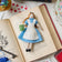 Hand-felted wool ornament of Alice from Alice in Wonderland, wearing her classic blue and white dress and holding a felt copy of Lewis Carroll's book. Crafted with embroidered details and made from merino-blend wool, the ornament includes a hanging string and measures approximately 5.75 inches tall