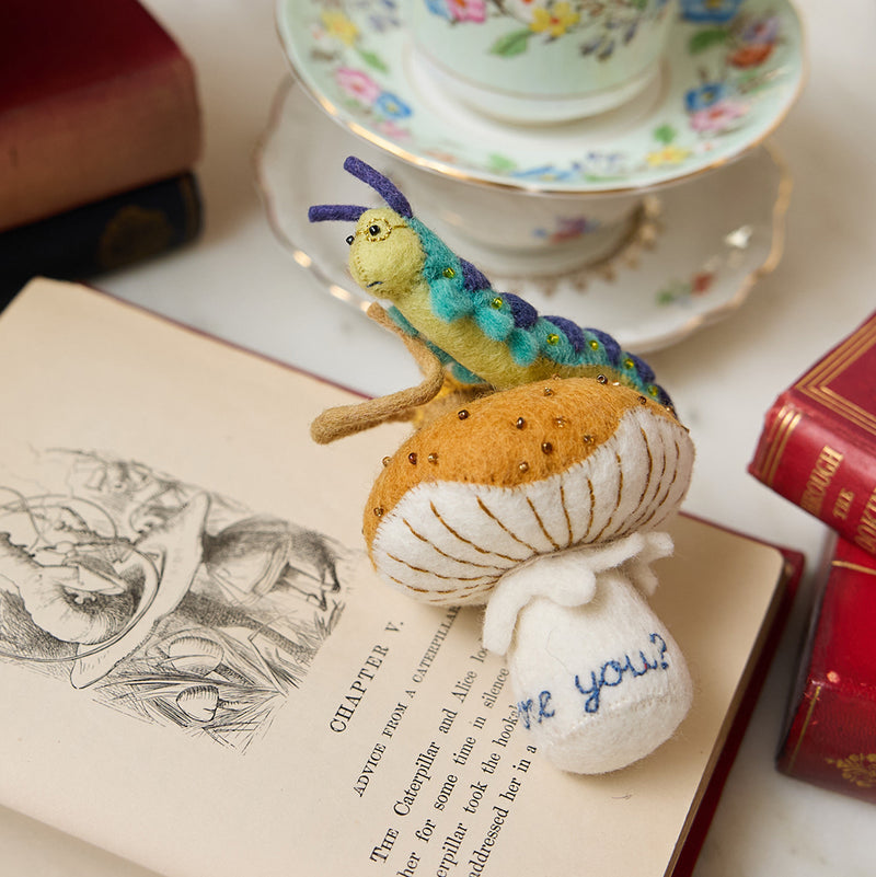 Hand-felted wool ornament of the Caterpillar from Alice in Wonderland, perched on a red-and-white mushroom with a hookah beside him. Features detailed embroidery and the phrase 'Who are you?' stitched at the base. Made from natural merino-blend wool, this whimsical ornament includes a hanging string and measures approximately 5.5 inches tall.