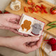 Miniature felt ornament shaped like a cookbook, hand-felted and embroidered with cooking-themed details including salt, pepper, vegetables, and a stovetop. Crafted from merino-blend wool with soft pages and a hanging string, this ornament measures approximately 2.5 inches tall. A cozy tribute to home cooking and culinary tradition. Open to page with a chicken and a boiling pot.