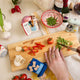 Miniature felt ornament shaped like a cookbook, hand-felted and embroidered with cooking-themed details including salt, pepper, vegetables, and a stovetop. Crafted from merino-blend wool with soft pages and a hanging string, this ornament measures approximately 2.5 inches tall. A cozy tribute to home cooking and culinary tradition.