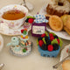 Hand-felted wool ornament featuring a pastel-blue teapot, matching teacup, and a tiered tower of colorful macarons.  Featured next to a berry pint ornament and mixed berry jam jar ornament. Embellished with embroidered details and soft textures, this miniature tea set includes a hanging string and measures approximately 2.5 inches tall. A charming nod to classic afternoon tea and vintage elegance.